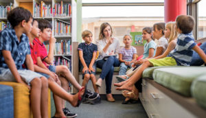 Children in discussion with teacher. All sitting on couches with books on shelves.